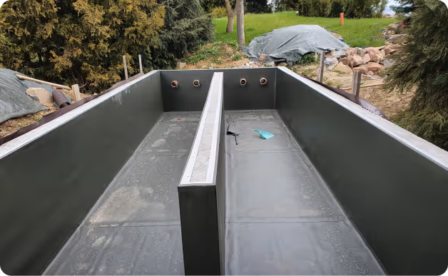 Leeres, rechteckiges Becken aus schwarzem Material im Freien mit einer Trennwand in der Mitte und umgeben von Bäumen und Gras.