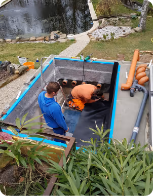 Zwei Männer verlegen Teichfolie in einem rechteckigen Teich im Bau neben einem Gartenteich.