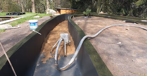 Baustelle mit einem langen, schwarzen Kunststoffbehälter im Freien, teilweise mit Sand gefüllt und mit Schläuchen darin, umgeben von Bäumen und einer Rasenfläche.