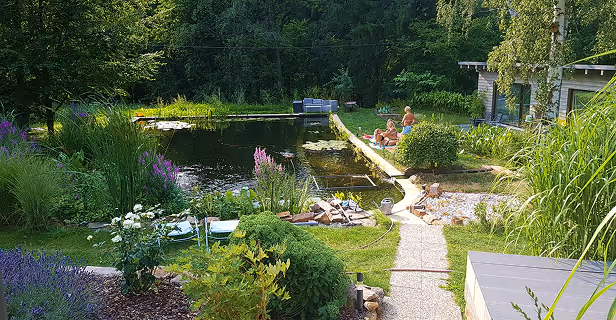 Ein natürlicher Gartenteich mit klarem Wasser, umgeben von Blumen, Sträuchern und Bäumen, mit Menschen, die am Ufer entspannen.